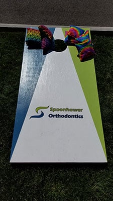 Cornhole game board featuring Spoonhower Orthodontics logo on green grass surface.