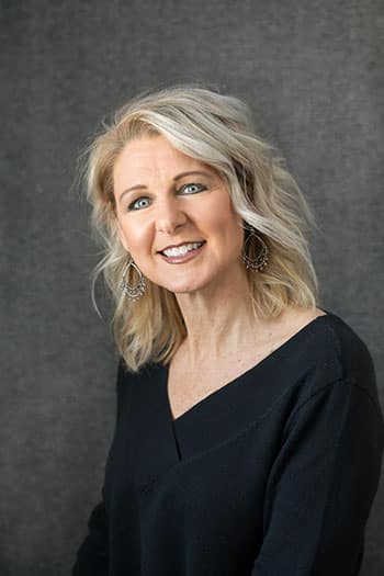 Professional headshot of a smiling woman with blonde hair wearing black against a gray background.
