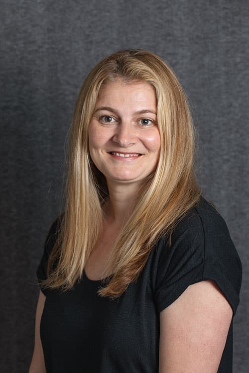 Professional headshot of a woman with long blonde hair smiling at camera against gray background.