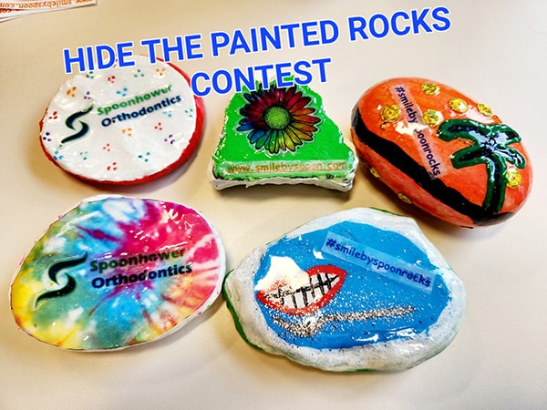 Five decorated painted rocks for "Hide the Painted Rocks Contest" by Spoonhower Orthodontics displayed on white surface.