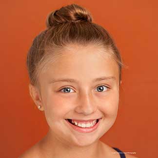 Young girl smiling at camera against orange background, wearing pearl earrings.
