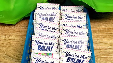 Stack of white and blue "You're the BALM!" orthodontic promotional cards on wooden surface with green fabric.