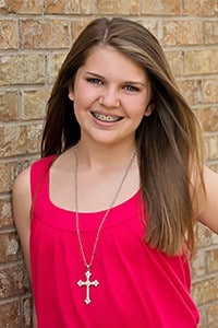 Young woman smiling at camera wearing pink tank top and cross necklace against brick wall.