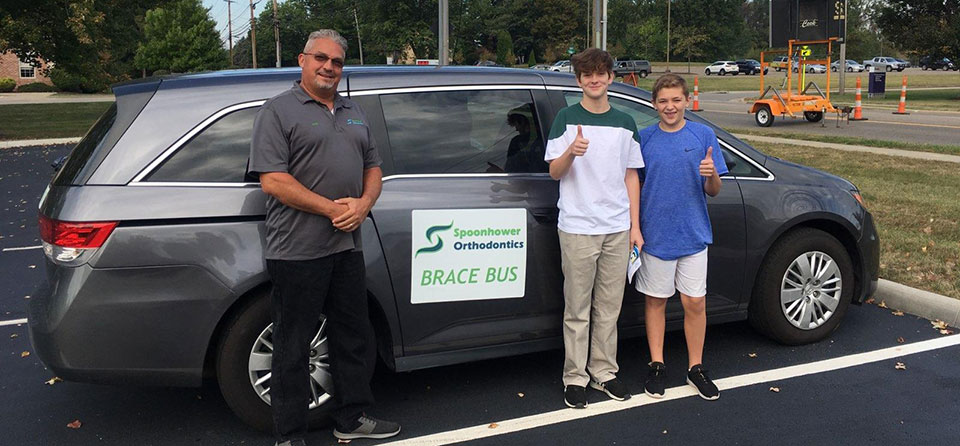 Men and boy stand beside two cars displaying Spoonhower Orthodontics signage at outdoor event.